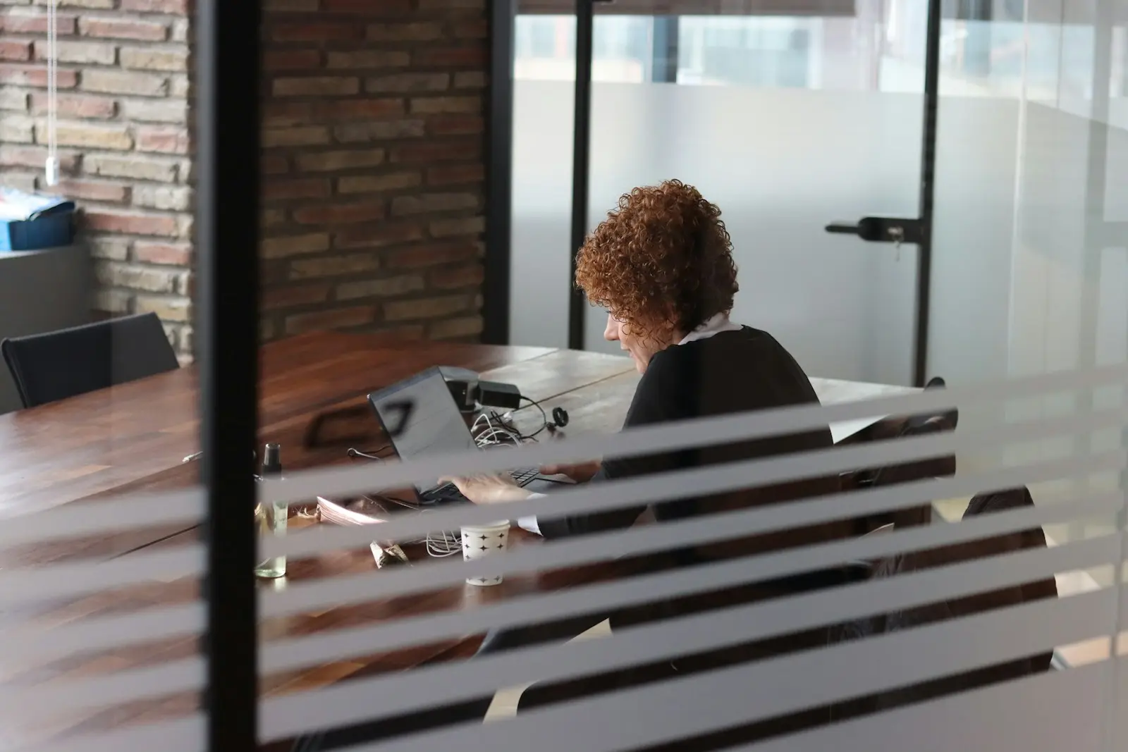 Un poste de travail aperçu derrière une cloison vitrée à bandes horizontales, dans un bureau aux murs de brique