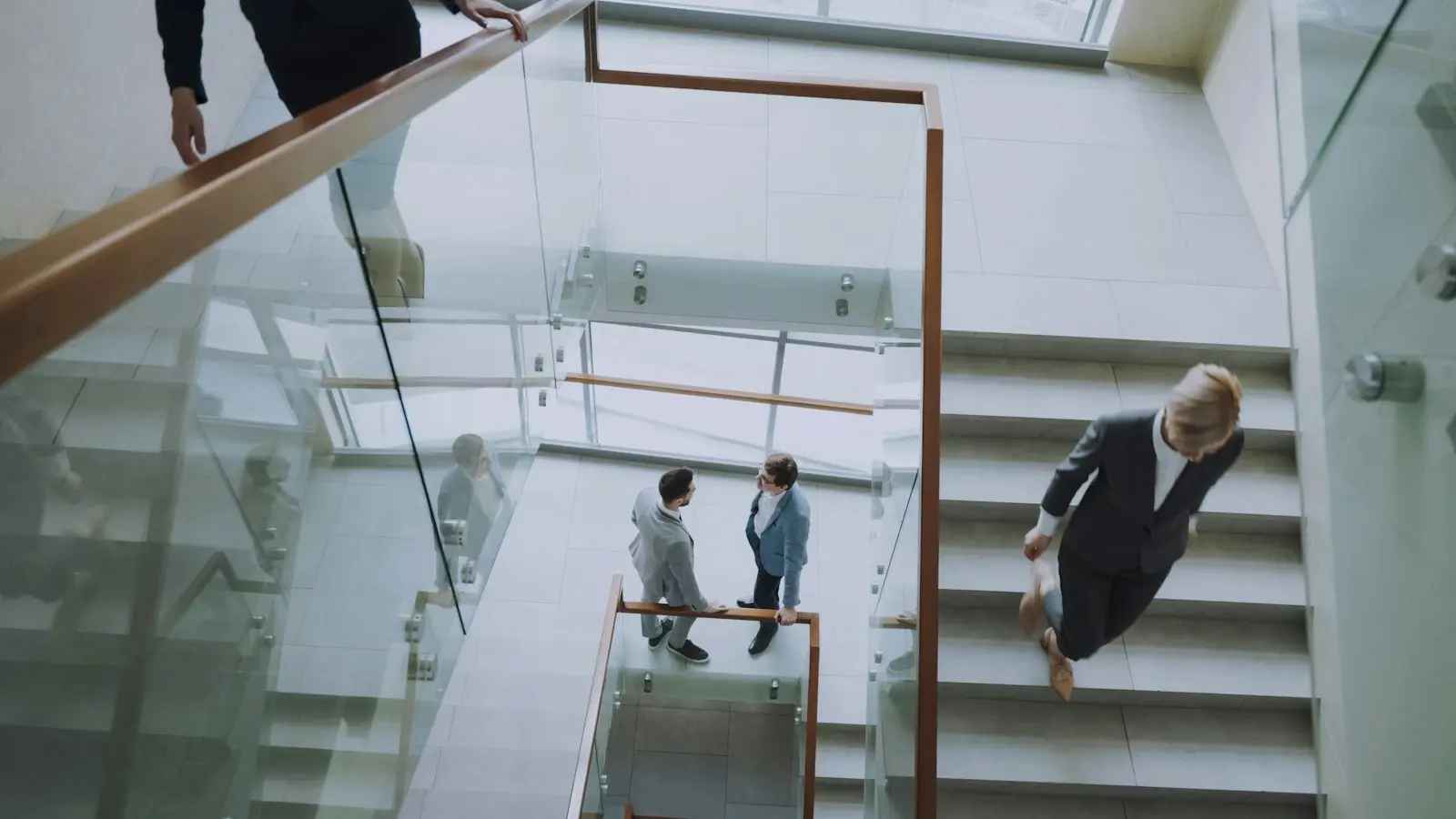 Deux personnes montent un escalier clair à rambarde vitrée dans un immeuble de bureaux