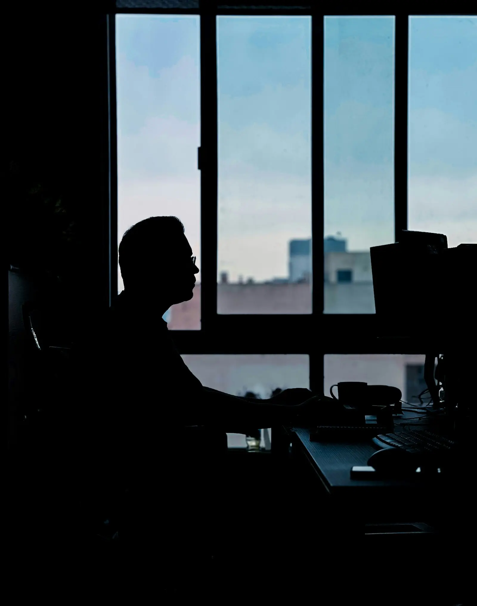 Silhouette d'une personne devant ses écrans, à contre-jour d'une fenêtre au crépuscule
